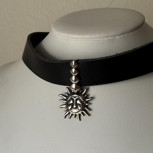 Può includere: Un collare choker in pelle nera con un ciondolo a forma di sole argentato e borchie piramidali argentate.
