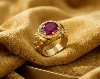 May include: A gold ring featuring a large, oval, deep red gemstone. The ring has a textured band and is set on a gold surface and a gold fabric. The ring is a statement piece.