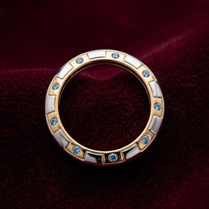 May include: A two-tone ring with a gold interior and silver exterior, accented with small blue gemstones. The ring is set against a dark red velvet background, highlighting its design and color.