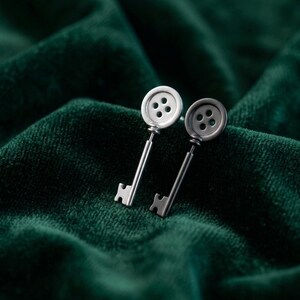 May include: A pair of silver-toned key-shaped earrings. Each earring features a button-like top with four holes and a slender key shaft. The earrings are displayed against a dark green, textured background.