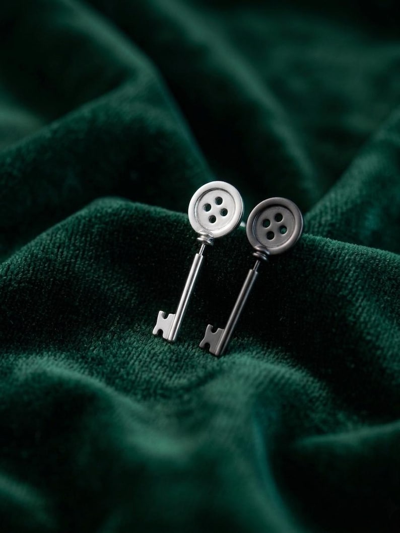 May include: A pair of silver-toned key-shaped earrings. Each earring features a button-like top with four holes and a slender key shaft. The earrings are displayed against a dark green, textured background.