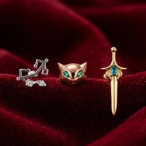 May include: Three pieces of jewelry: a silver earring with a musical note design, a gold cat-shaped earring with green eyes, and a gold sword-shaped earring with a blue gem. The jewelry is displayed on a red velvet surface.