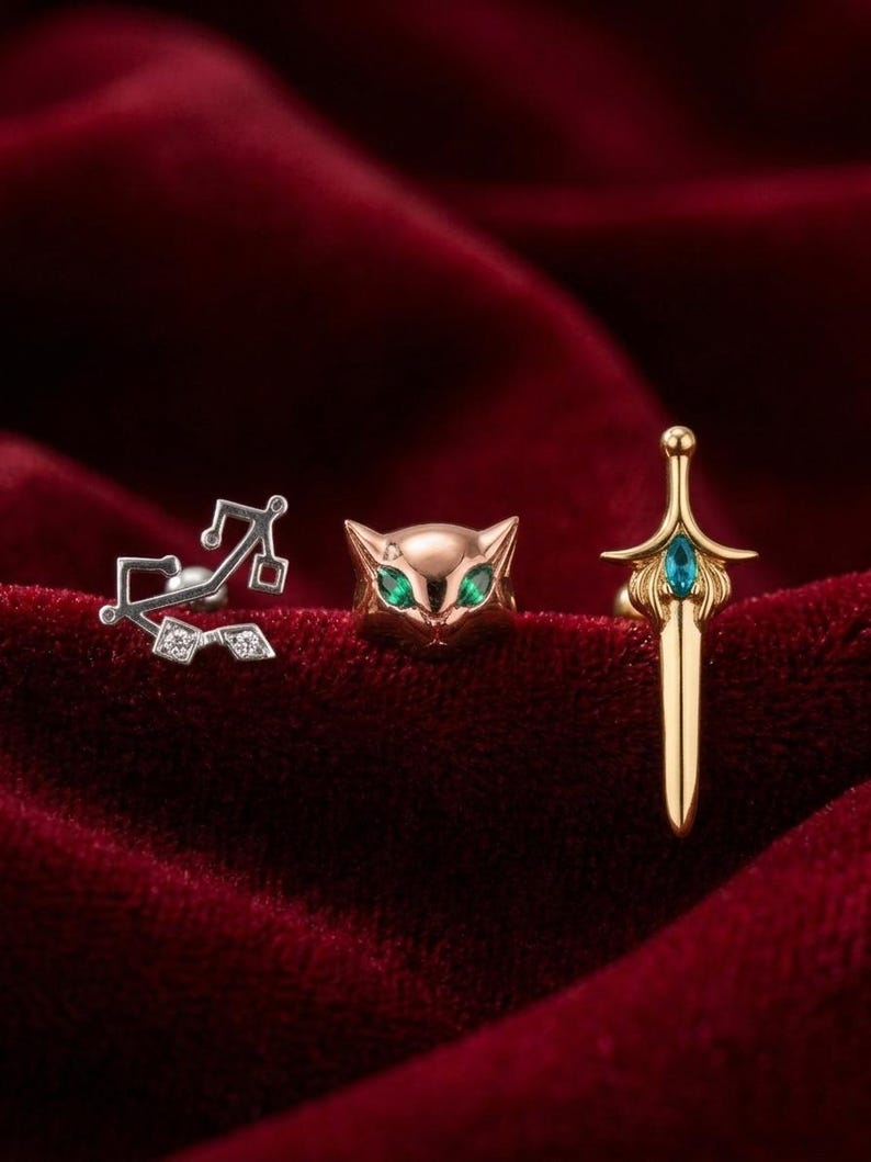 May include: Three pieces of jewelry: a silver earring with a musical note design, a gold cat-shaped earring with green eyes, and a gold sword-shaped earring with a blue gem. The jewelry is displayed on a red velvet surface.