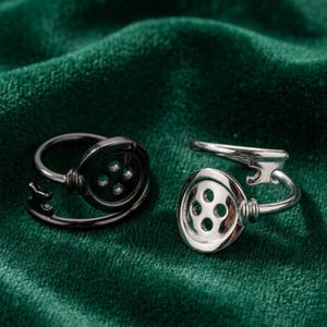 Peut inclure: Deux bagues avec des motifs en forme de bouton. Une bague est noire, l'autre est argentée. Chaque bague a une face de bouton avec quatre trous et une bande incurvée. Les bagues sont posées sur un fond de velours vert foncé.