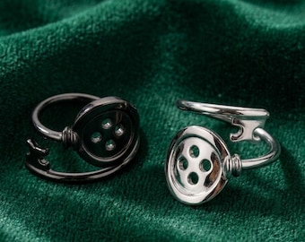 Puede incluir: Dos anillos con diseños en forma de botón. Un anillo es negro, el otro plateado. Cada anillo tiene una cara de botón con cuatro agujeros y una banda curva. Los anillos están sobre un fondo de terciopelo verde oscuro.