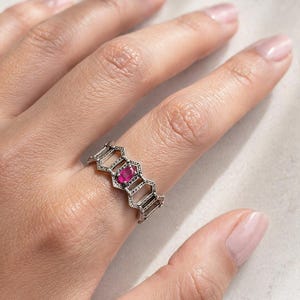 May include: Silver ring with a honeycomb design, featuring a vibrant pink gemstone at its centre. The ring is worn on a finger, showcasing its intricate openwork pattern and the gemstone's colour.
