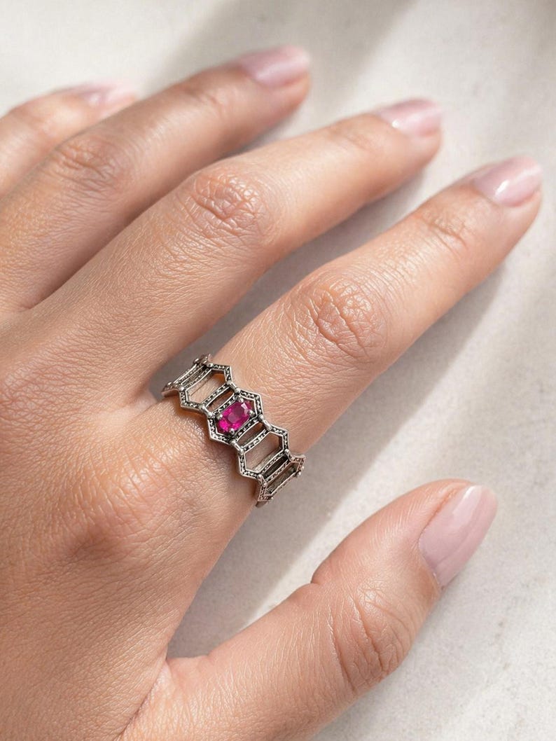May include: Silver ring with a honeycomb design, featuring a vibrant pink gemstone at its centre. The ring is worn on a finger, showcasing its intricate openwork pattern and the gemstone's colour.
