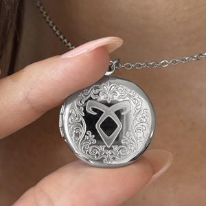 May include: A circular silver locket pendant suspended from a matching silver chain. The front of the locket features an intricate engraved filigree border surrounding a central embossed symbol that resembles a stylized letter A with a loop.