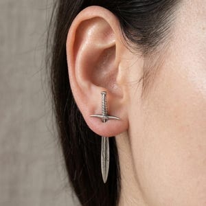May include: A silver-colored sword earring. The earring is a miniature replica of a sword, with a detailed hilt and blade. The earring is worn in the ear, with the blade hanging down. The background is a neutral gray.