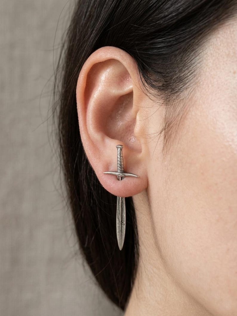 May include: A silver-colored sword earring. The earring is a miniature replica of a sword, with a detailed hilt and blade. The earring is worn in the ear, with the blade hanging down. The background is a neutral gray.