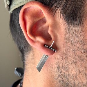 May include: A silver earring with a small, rectangular bar and a dangling knife-shaped charm. The charm has a small, clear stone set in it.