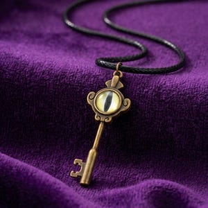 May include: A bronze-colored key pendant with an eye-shaped center, suspended from a black cord necklace. The eye has a yellow iris and a black pupil. The key has ornate detailing and is set against a deep purple background.