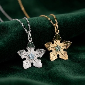 Może przedstawiać: Dwa naszyjniki z wisiorkami w kształcie liści. Jeden jest posrebrzany, drugi pozłacany. Każdy wisiorek ma mały, jasnoniebieski kamień szlachetny pośrodku. Naszyjniki spoczywają na ciemnozielonej aksamitnej powierzchni.