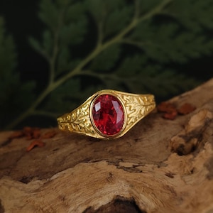 Op de afbeelding: Een gouden ring met een ovaal geslepen, felrode edelsteen in het midden. De ringband is voorzien van een gedetailleerd, verhoogd bladpatroon dat zich aan beide zijden van de zetting van de steen uitstrekt, wat zorgt voor een textuur.