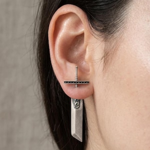 May include: A silver earring with a sword design. The earring has a rectangular blade shape and a crossbar with small black details. The earring is worn on the ear, with the post visible.