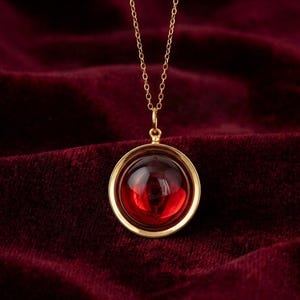 May include: A gold-colored necklace with a pendant featuring a large, round, red gemstone. The gemstone is set in a gold bezel and hangs from a delicate gold chain. The background is a deep red velvet.