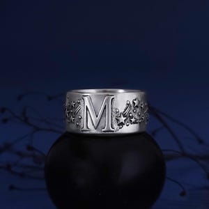 May include: A silver-toned metal ring featuring a prominent capital letter M embossed on the front. The band is decorated with a celestial pattern of various sized stars and shooting star motifs that wrap around the surface of the ring.