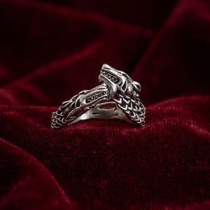 May include: A silver-colored ring featuring a detailed wolf head design. The ring showcases intricate carvings of the wolf's fur and facial features. The ring is set against a deep red velvet background.