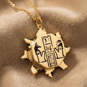 Può includere: Collana con pendente dorato con un design a tema egiziano. Il pendente a forma di stella presenta dettagli neri e simboli geroglifici. La collana ha una catena sottile.