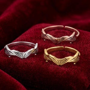 May include: Three adjustable rings in silver, rose gold, and gold tones. Each ring features a stylized wing design. The rings are displayed on a deep red velvet surface, creating a striking contrast.