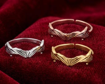 May include: Three adjustable rings in silver, rose gold, and gold tones. Each ring features a stylized wing design. The rings are displayed on a deep red velvet surface, creating a striking contrast.