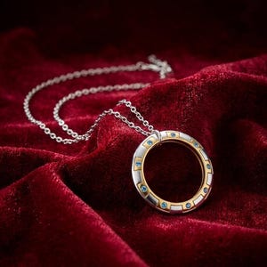 May include: A silver necklace with a circular pendant. The pendant has a gold and silver design with small blue gemstones. The necklace is displayed on a red velvet surface.