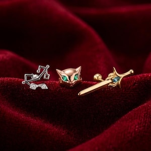May include: Three stud earrings on a red velvet surface. One is a silver musical note design with small clear stones. The second is a gold cat face with green eyes. The third is a gold sword with blue accents.