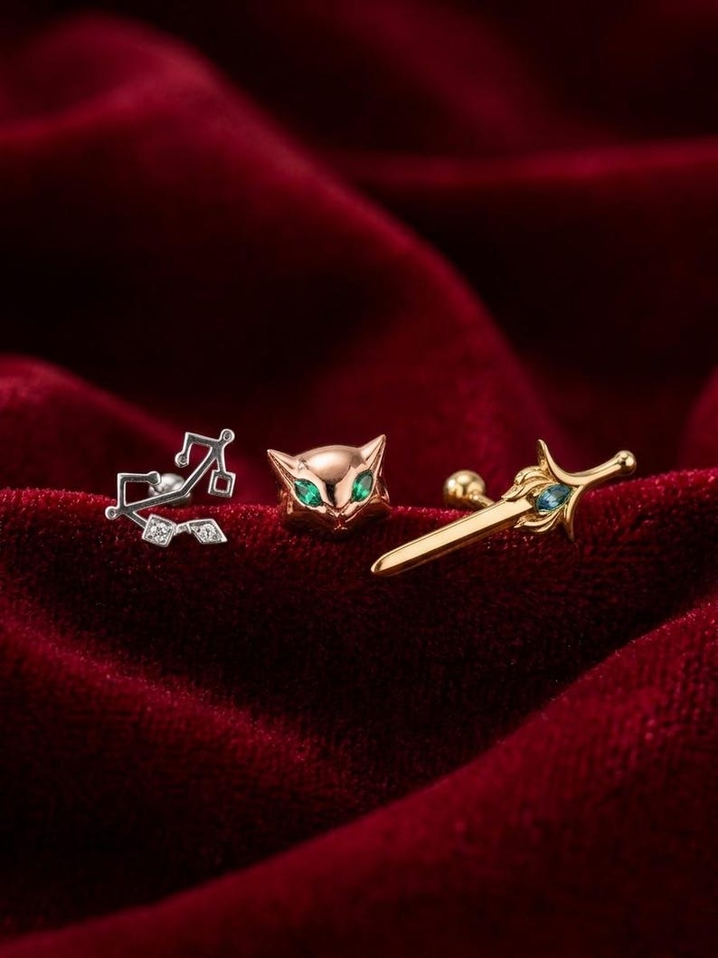 May include: Three stud earrings on a red velvet surface. One is a silver musical note design with small clear stones. The second is a gold cat face with green eyes. The third is a gold sword with blue accents.