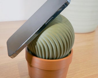 May include: A black smartphone rests on top of a green, textured, spherical object. The object is in a brown terracotta pot.