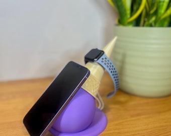 May include: A purple phone stand shaped like an ice cream cone holds a black smartphone and a blue smartwatch.