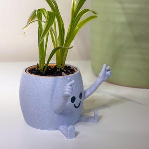 May include: A light blue, speckled ceramic planter with a cartoon face and arms, holding a small green and white plant. The planter has a smiling face and is sitting with its legs extended. Another green planter is in the background.