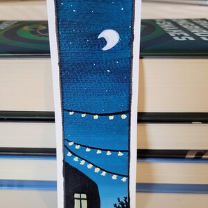 May include: A hand-painted bookmark featuring a night scene with a crescent moon, stars, a silhouette of a house with a window, and fairy lights.