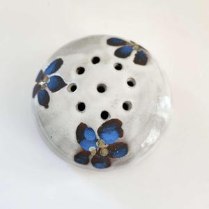 May include: A white ceramic flower pot with blue and brown flower designs. The pot has a round shape and multiple holes on the top.