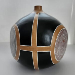 May include: A black gourd-shaped vase with a brown neck and decorative tan and white geometric designs. The vase has a rounded body with a cross-like pattern and circular designs on the sides. The vase is a decorative home decor item.