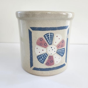May include: A beige ceramic crock with a blue square and a floral design. The floral design has white, red, and blue elements. The crock has a rounded top and a cylindrical shape, suitable for kitchen storage.