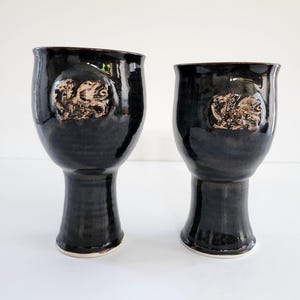 May include: Two black ceramic goblets with a textured, raised design on the front. The goblets have a wide rim and a pedestal base. The design on the front appears to be a relief of a mythical creature. The interior of the goblets is a dark brown colour.