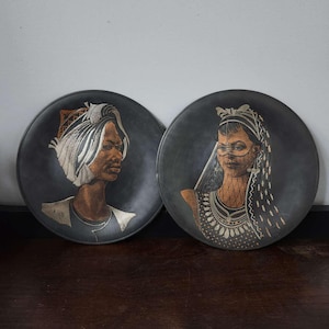 May include: Two round decorative plates with portraits of people wearing head coverings. The plates have a dark gray background with detailed illustrations in shades of brown, white, and gold. The plates are displayed on a dark wooden surface.