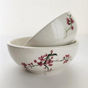 May include: Two white ceramic bowls, one stacked atop the other. Each bowl features a delicate floral design with pink blossoms and green stems. The bowls are likely for serving food.
