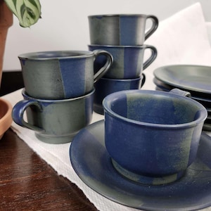 May include: A set of blue and green ceramic coffee cups and saucers. The cups have a striped design and are stacked on top of each other. The saucers are also striped and have a slightly raised edge.