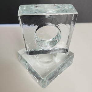Peut inclure: Deux bougeoirs carrés en verre transparent. Chaque support comporte une ouverture circulaire au centre. Le verre a une surface texturée et les supports sont empilés l'un sur l'autre.