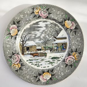 May include: A decorative plate with a winter scene and floral border. The plate has a grey background with a winter landscape in the centre and rose clusters around the edge. The roses are pink, yellow, and white.