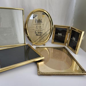 May include: A selection of gold-coloured brass picture frames in various shapes and sizes. The frames include rectangular, oval, and folding designs. Some frames have glass and backing, while others are empty. The frames are arranged on a white surface.