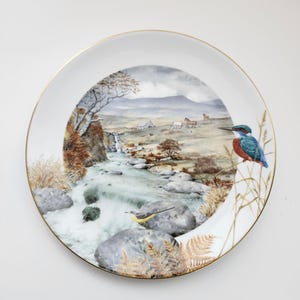 May include: A decorative white plate with a gold rim featuring a landscape scene with a river, trees, and horses. A blue and orange bird is perched on the edge of the plate. The plate has a detailed, painted design.