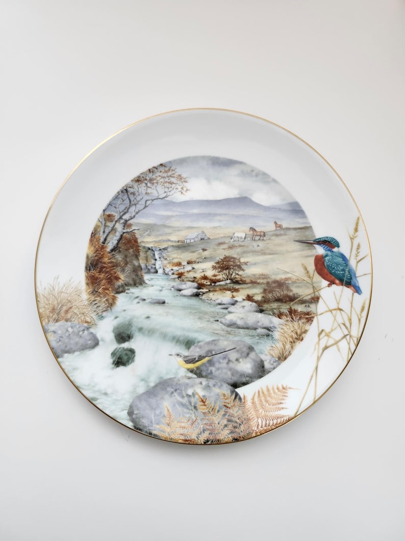 May include: A decorative white plate with a gold rim featuring a landscape scene with a river, trees, and horses. A blue and orange bird is perched on the edge of the plate. The plate has a detailed, painted design.