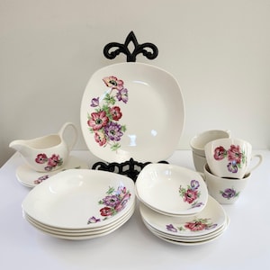 May include: A vintage white porcelain dinnerware set featuring a floral design with red, purple, and pink flowers. The set includes plates, bowls, cups, saucers, and a gravy boat. A black decorative stand is behind the large plate.