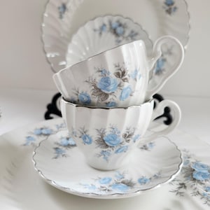 May include: A vintage white porcelain tea set featuring a blue rose design and silver edging. The set includes two teacups, saucers, and a larger plate, all with a fluted edge. The teacups are stacked.