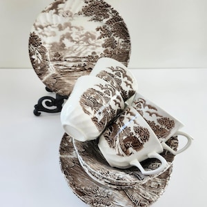 May include: A vintage tea set featuring brown and white ceramic teacups and saucers with a landscape design. The set includes two teacups, two saucers, and a large plate, all with a matching pattern. The teacups are white with a brown landscape design.