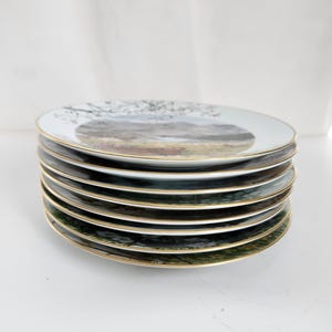 May include: A stack of eight white ceramic plates with a gold rim. Each plate features a landscape scene with trees, mountains, and a field. The plates are stacked on top of each other.