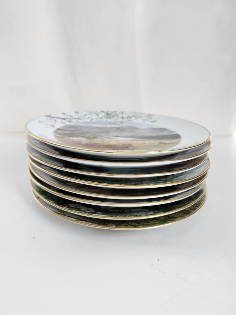 May include: A stack of eight white ceramic plates with a gold rim. Each plate features a landscape scene with trees, mountains, and a field. The plates are stacked on top of each other.