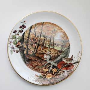 May include: A decorative white plate with a gold rim featuring a detailed forest scene. The scene includes a squirrel, mushrooms, a bird, and butterflies. The plate also has a branch with blackberries.